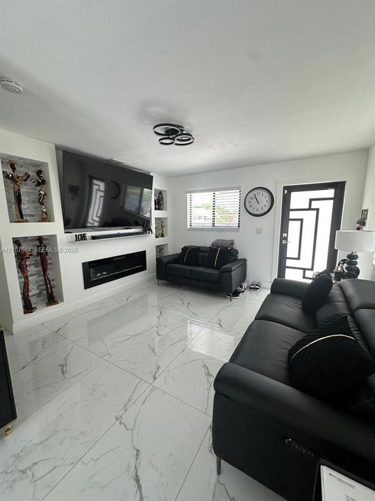 Large-format porcelain tile flooring, built-in wall niches, linear electric fireplace, modern flush-mount multi-ring ceiling lighting, and geometric-patterned glass entry door