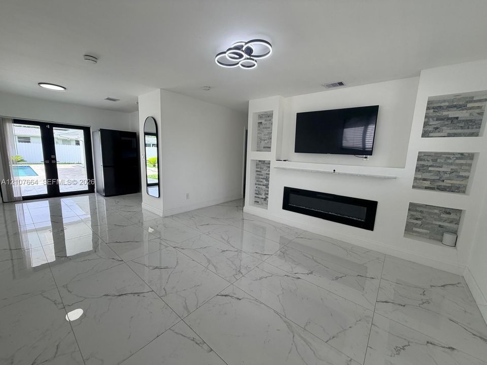 Large-format tile flooring, built-in electric fireplace, integrated shelving with stone-finish accents, modern circular LED light fixture, and double glass-paned doors