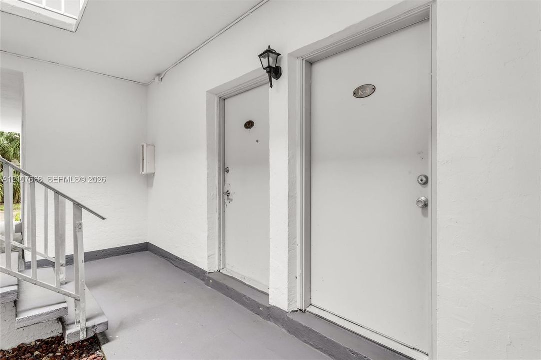 Covered entryway featuring white exterior walls, painted concrete flooring, a metal staircase with railing, and wall-mounted exterior lighting
