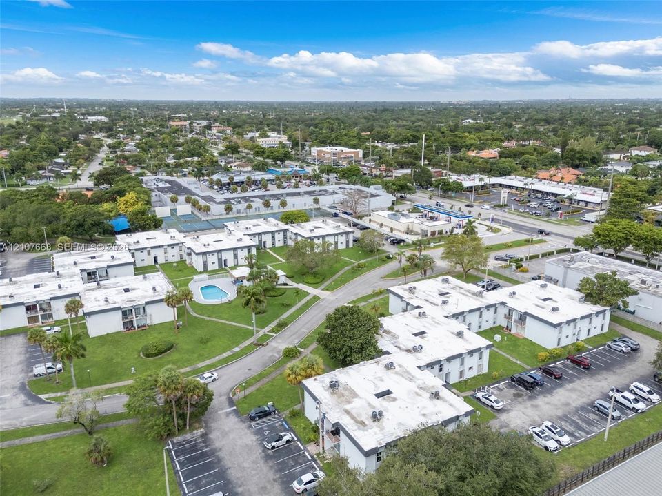 Aerial perspective of a multi-unit complex featuring an outdoor swimming pool, paved surface parking areas, manicured green space, and mature palm trees