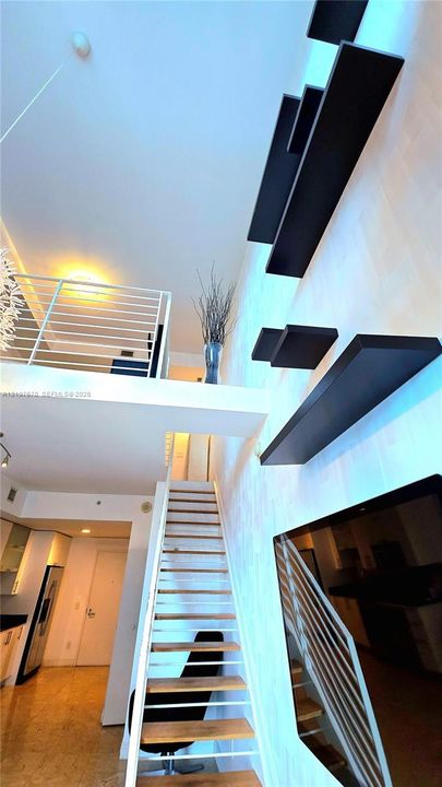 Double-height ceiling, floating staircase with open wood-finish treads, metal horizontal-bar railings, and built-in staggered wall shelving