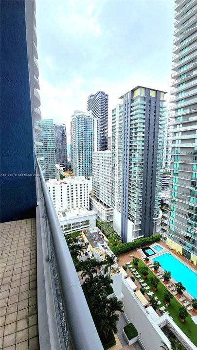 Private balcony featuring tiled flooring, a metal railing, and a cityscape backdrop with access to a swimming pool and landscaped grounds