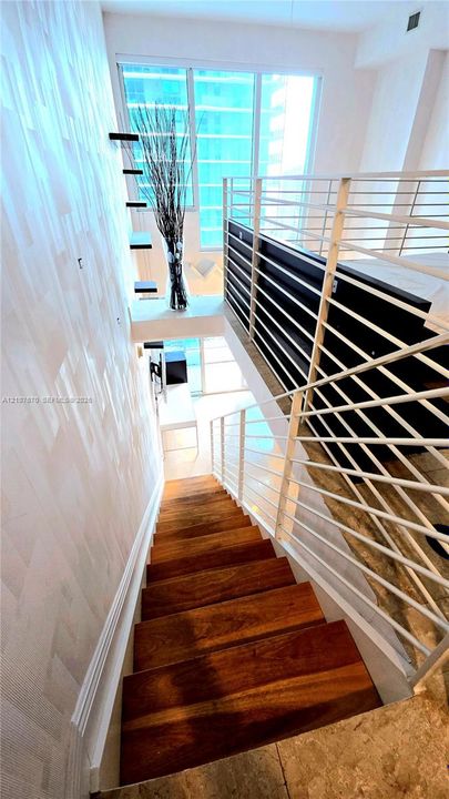 Staircase with wood-finish treads, floor-to-ceiling windows, white horizontal metal railing, and textured wall finish