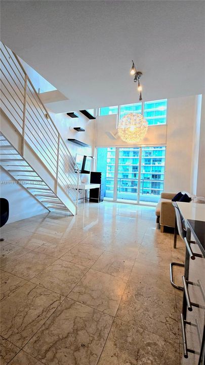 Open-concept living area featuring stone tile flooring, double-height ceilings, floor-to-ceiling windows, a glass-panel balcony door, and a floating metal staircase with integrated railing