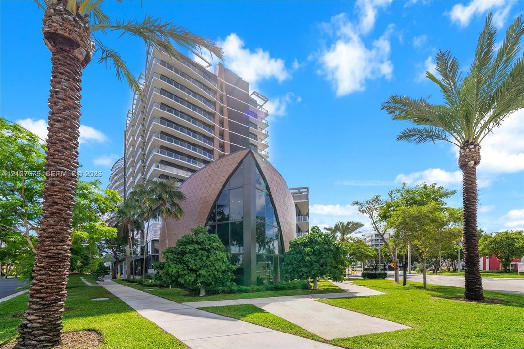 High-rise residential exterior featuring expansive glass curtain walls, private balconies, a unique arched architectural structure, and manicured grounds with palm trees