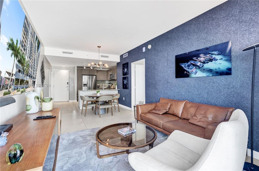 Open-concept living space featuring large-format tile flooring, a prominent textured navy accent wall, stainless steel kitchen appliances, and a contemporary multi-light chandelier