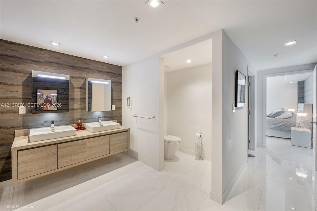 Floating dual-sink vanity with light wood-finish cabinetry, white rectangular vessel sinks, textured wood-panel accent wall, and polished stone-finish floor tiling