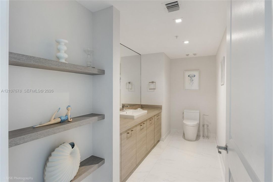 Bathroom featuring built-in wood-finish shelving, vanity with a rectangular vessel sink, large wall-mounted mirror, recessed ceiling lighting, and large-format tile flooring