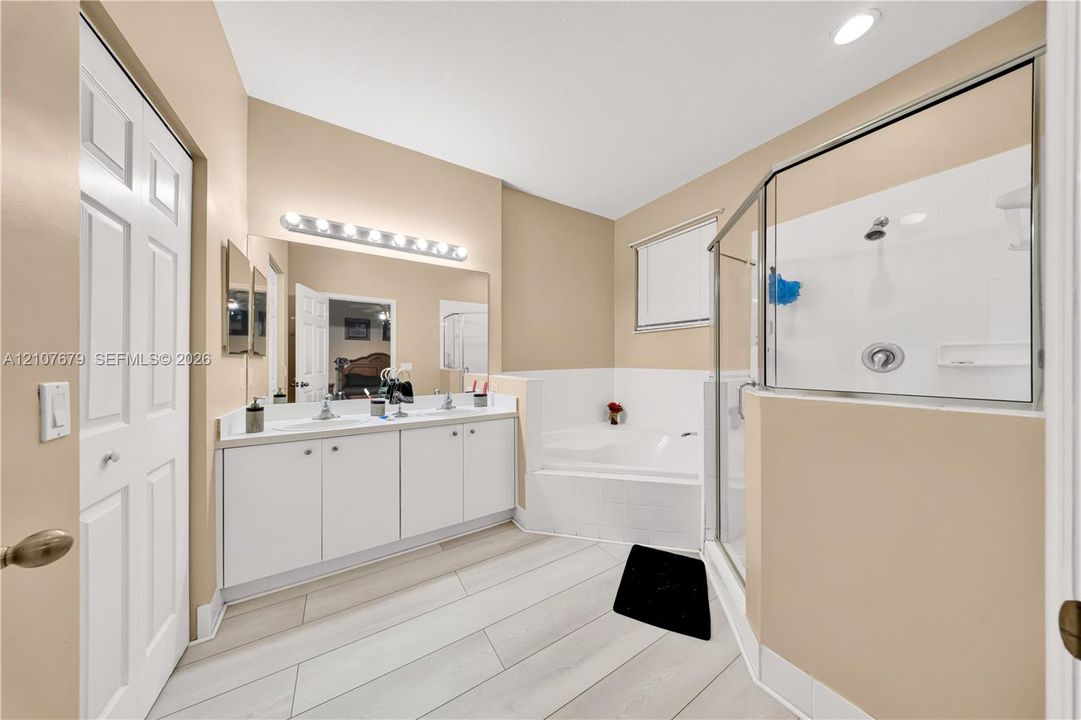 Dual-sink vanity with white cabinetry, rectangular drop-in sinks, wall-to-wall mirror, tiled soaking tub, and enclosed glass shower with white surround