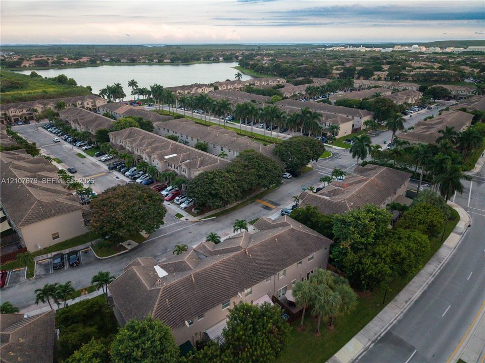 Aerial perspective of a multi-unit residential community featuring clay-tile roofing, established palm trees, paved thoroughfares, and proximity to a large body of water