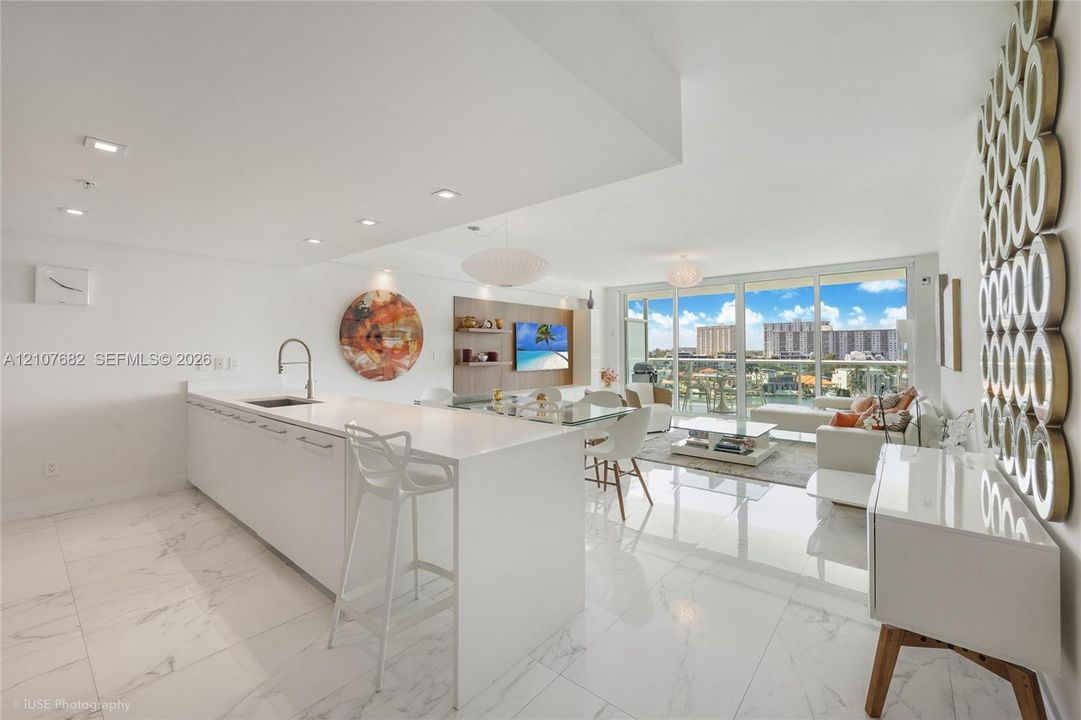 Open-concept interior featuring large-format tile flooring, a kitchen island with stone-finish countertops, recessed lighting, and floor-to-ceiling glass sliding doors leading to a balcony