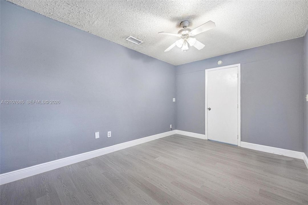 Neutral-toned interior room featuring wood-finish flooring, a ceiling-mounted light fixture with fan blades, and a solid-core interior door