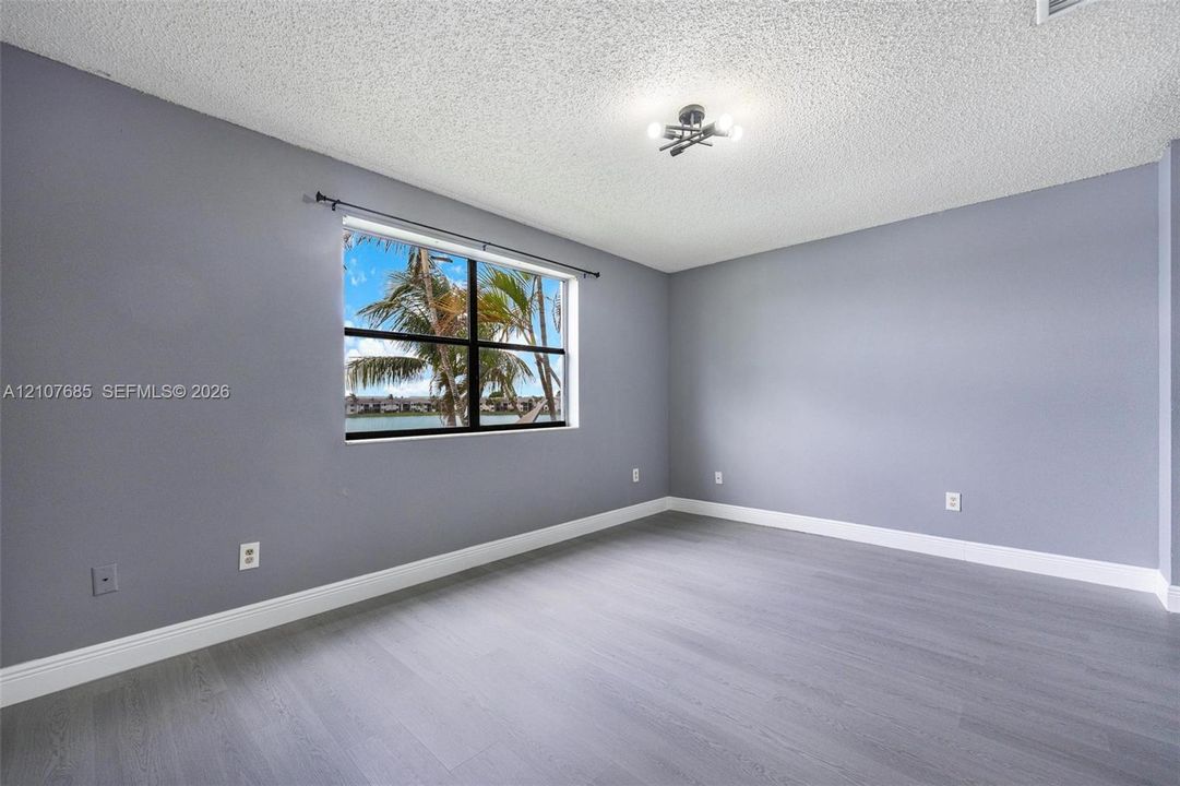 Room featuring gray-tone walls, wood-finish flooring, a multi-bulb flush-mount light fixture, and a single-pane window with exterior water and palm tree scenery