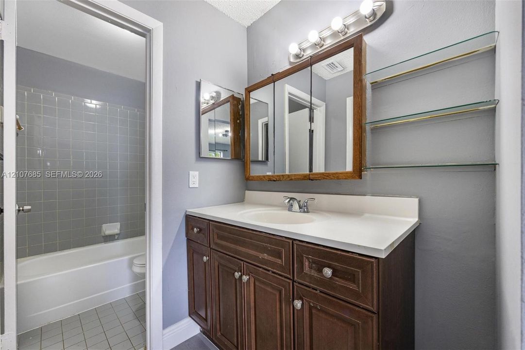 Bathroom featuring a vanity with wood cabinetry, integrated sink, triple-mirror medicine cabinet, four-light vanity fixture, and glass shelving