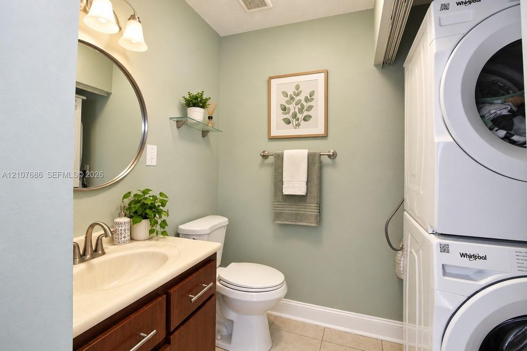 Laundry area featuring a stackable washer and dryer, wood-finish vanity with an integrated basin, circular wall mirror, and glass shelving