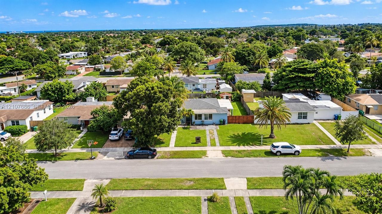 Aerial perspective of a single-story residence featuring a front lawn, mature canopy trees, a concrete driveway, and palm landscaping