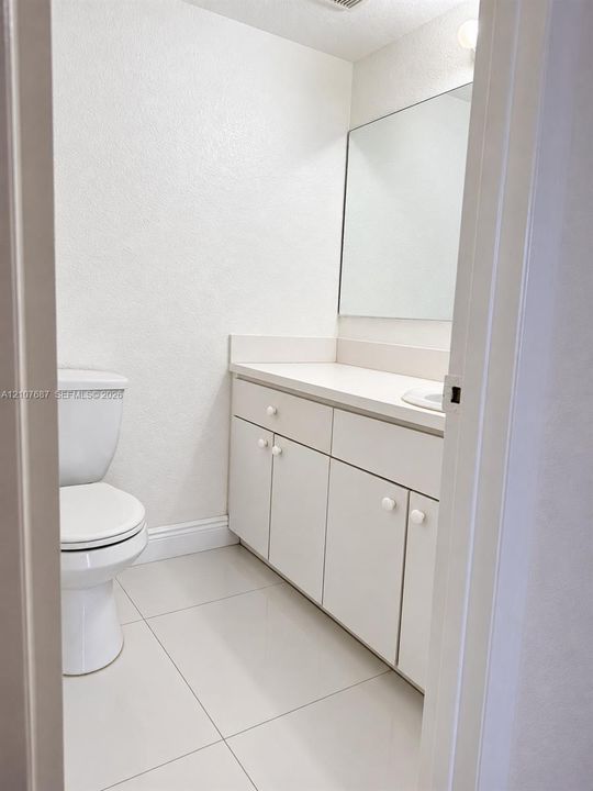 Bathroom featuring white cabinetry, solid-surface countertop with integrated sink, large wall-mounted mirror, white ceramic tile flooring, and standard toilet
