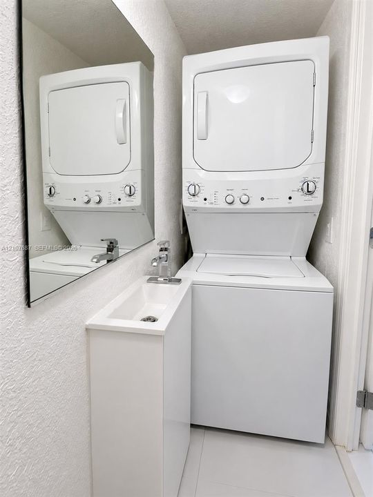 Stacked laundry unit, wall-mounted mirror, white pedestal-style sink, and chrome single-handle faucet