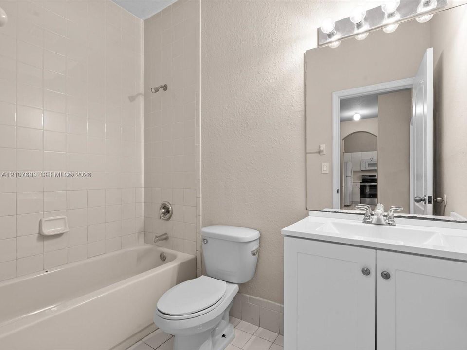 Bathroom featuring a white vanity cabinet with a integrated sink, expansive wall-mounted mirror, multi-bulb vanity light, and a tiled shower surround