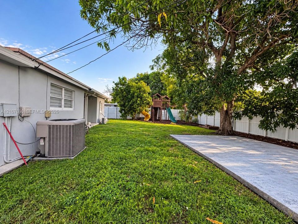 Spacious backyard featuring a rectangular concrete patio, mature trees, white vinyl privacy fencing, and exterior stucco siding