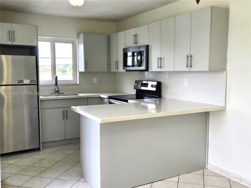 Kitchen featuring flat-panel cabinetry with metal hardware, light-toned solid surface countertops, a stainless steel refrigerator, a built-in microwave, and ceramic tile flooring