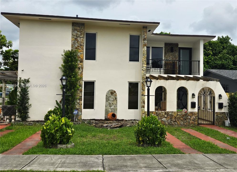 Two-story exterior featuring a stone-clad facade, arched entryway detailing, a covered second-story balcony with metal railing, and a pergola-style overhang