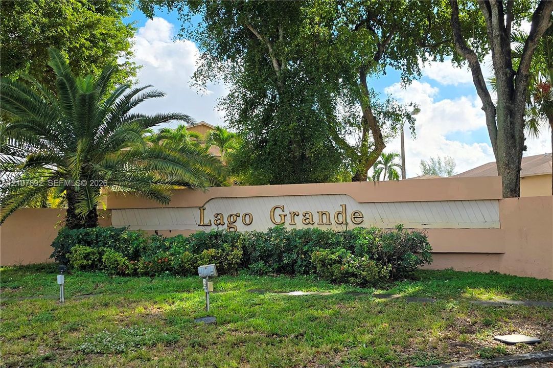 Lago Grande monument sign featuring metal lettering, stucco wall construction, mature palm trees, and manicured shrubbery