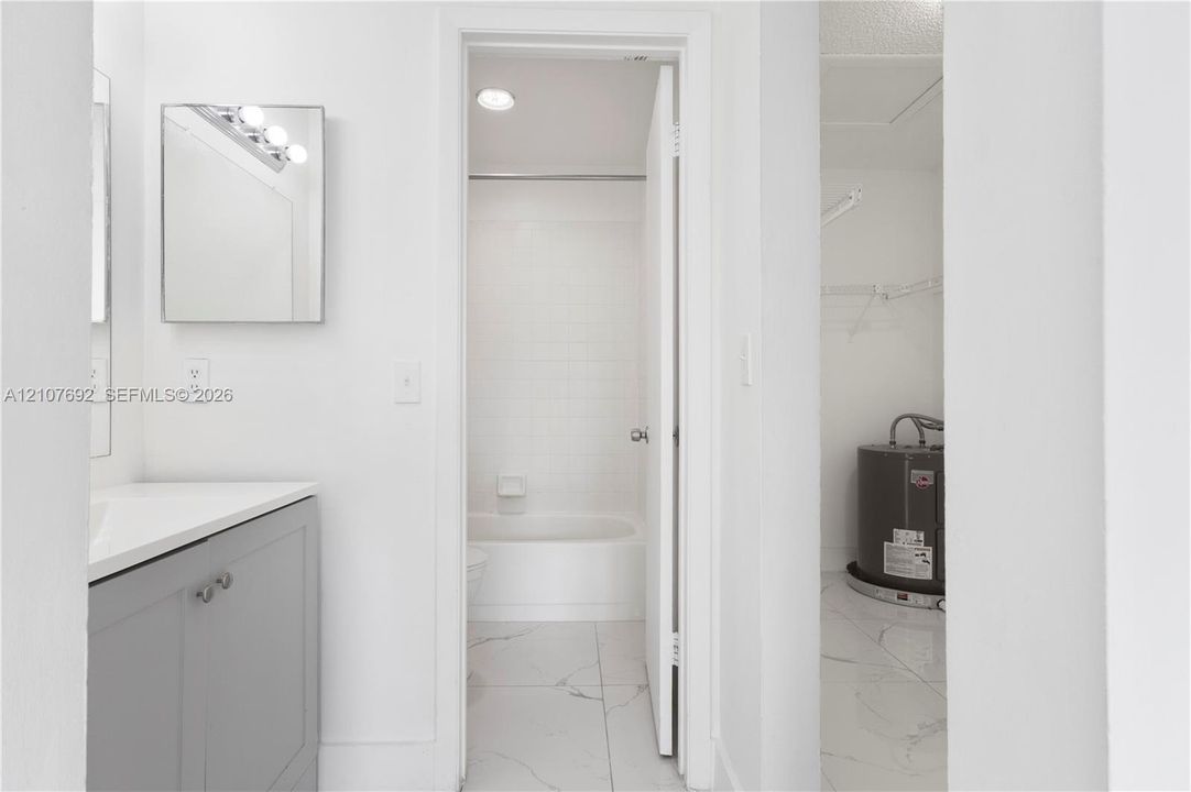 Bathroom featuring a grey vanity with a white countertop, wall-mounted mirror, light-colored tile flooring, and a tub with a tile surround