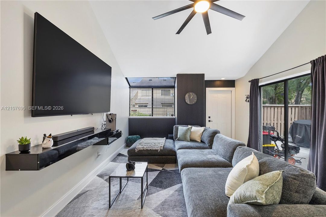 Vaulted ceilings, dark accent wall with vertical slat detailing, matte black ceiling fan with integrated lighting, floating black media console, and sliding glass doors