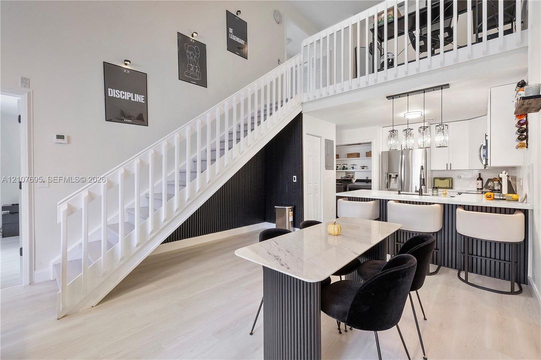 Double-height ceiling with an open overlook, staircase featuring vertical slat paneling, white shaker-style cabinetry, breakfast bar with pendant lighting, and light-toned wood-finish flooring
