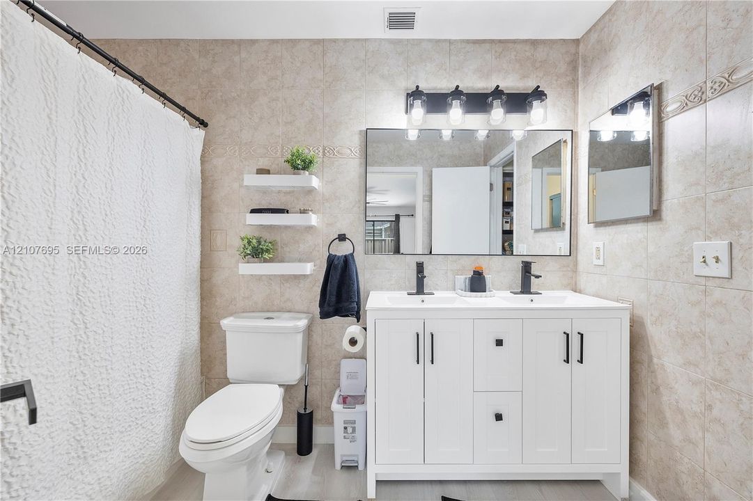 Double-sink vanity with white cabinetry, matte black hardware, and rectangular undermount sinks, complemented by a wide vanity mirror, four-light wall fixture, and neutral-toned wall tiling