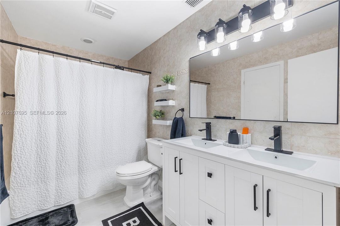 Dual-sink vanity with white cabinetry, matte black hardware, stone-surface countertop, and rectangular undermount sinks, complemented by a large-format mirror with integrated multi-bulb vanity lighting and neutral tile wall cladding