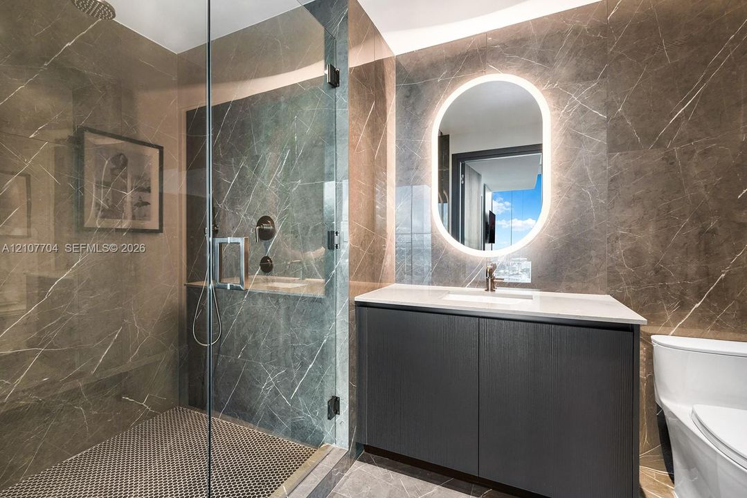 Bathroom featuring floor-to-ceiling stone tile, a glass-enclosed shower, a modern dark-finish vanity, and a backlit oval vanity mirror