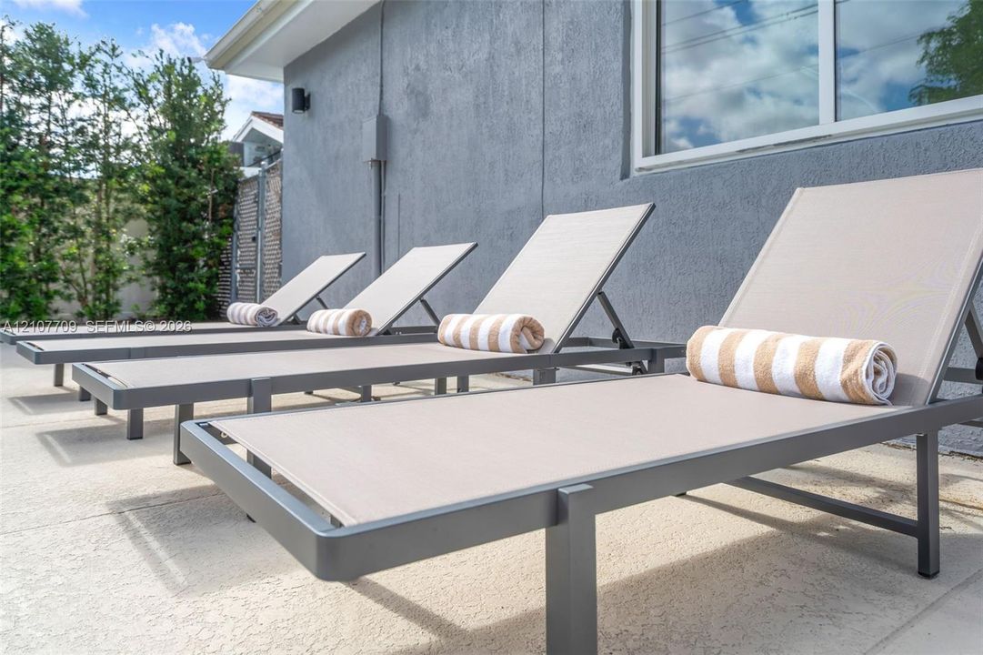Textured gray stucco exterior featuring a concrete patio surface, white-trimmed window, and tall perimeter hedges