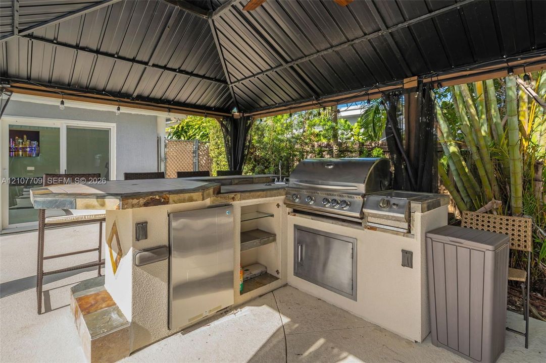 Outdoor kitchen featuring a stone-clad island with a counter, stainless steel built-in grill, stainless steel refrigerator, and protective metal-roofed gazebo