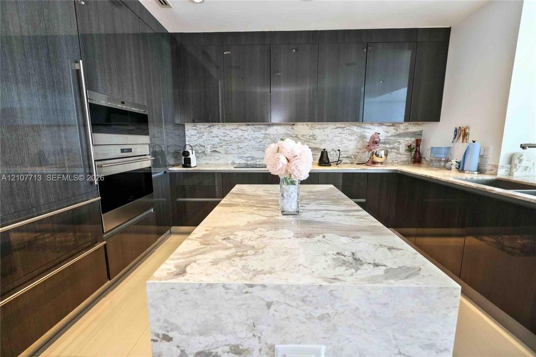 Modern kitchen featuring high-gloss cabinetry, stone countertops with waterfall-edge island, built-in stainless steel wall oven, and built-in microwave