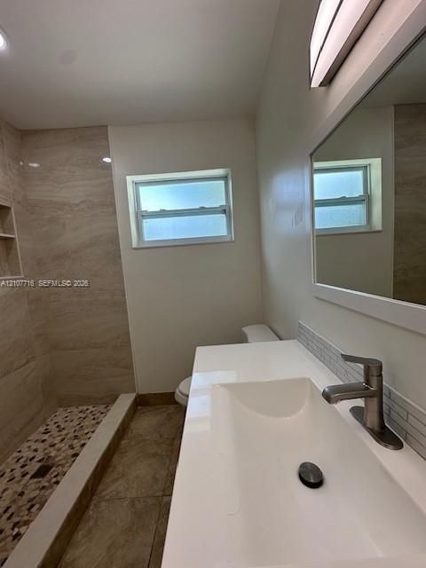 Bathroom featuring a white vanity with an integrated rectangular sink and metallic faucet, stone-tile flooring, and a walk-in shower with textured tile surround, pebble-style shower floor, and built-in shelving