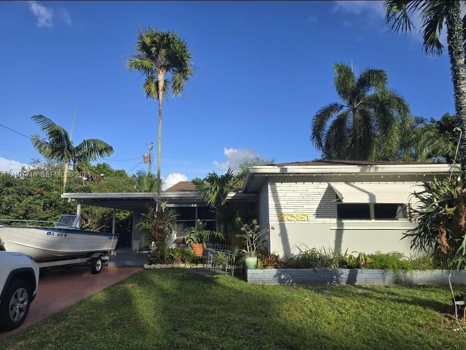 Single-story residence featuring a painted masonry facade, metal awning window coverings, a covered carport, and established tropical landscaping