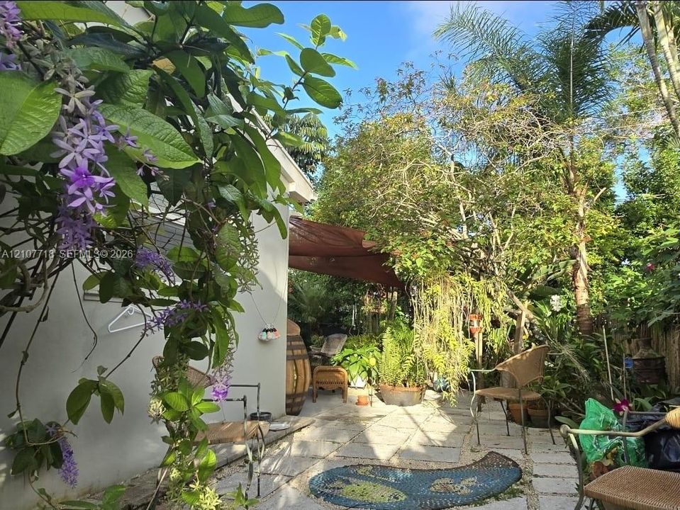 Landscaped outdoor area featuring stone paver flooring, established trees, flowering vines, and a textile shade canopy