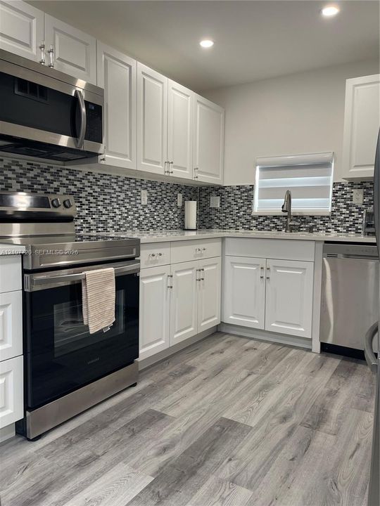 Kitchen featuring white shaker-style cabinetry, stainless steel appliances, multi-tonal mosaic tile backsplash, stone-finish countertops, and wood-finish flooring