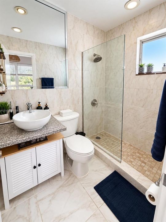 Bathroom featuring a vessel sink with a stone-finish countertop, white louvered vanity cabinetry, large wall-mounted mirror, walk-in shower with a frameless glass enclosure, and tiled flooring