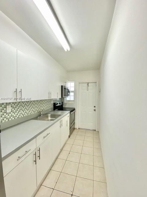 Kitchen featuring white cabinetry with brushed metal hardware, solid-surface countertops, mosaic tile backsplash, integrated stainless steel sink, and ceramic tile flooring