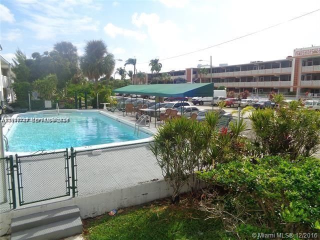 Outdoor swimming pool featuring a concrete deck, a metal perimeter fence with a gate, a covered awning structure, and mature landscaping