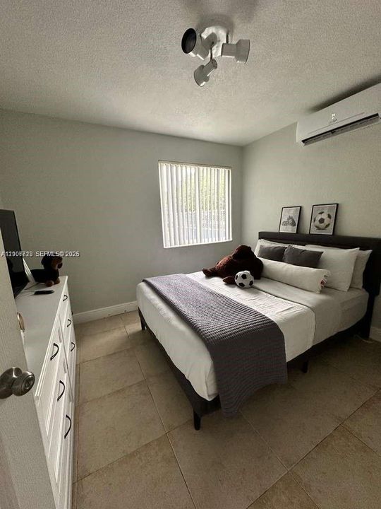 Room interior featuring large format tile flooring, wall-mounted mini-split air conditioning unit, triple-head directional ceiling light fixture, and vertical blinds