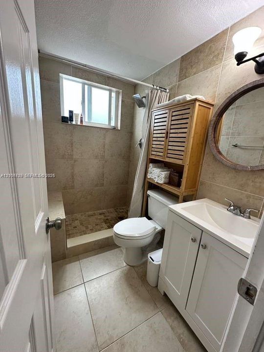 Bathroom featuring stone-finish wall and floor tile, walk-in shower with integrated bench, white vanity cabinet with a ceramic sink, and a wall-mounted light fixture