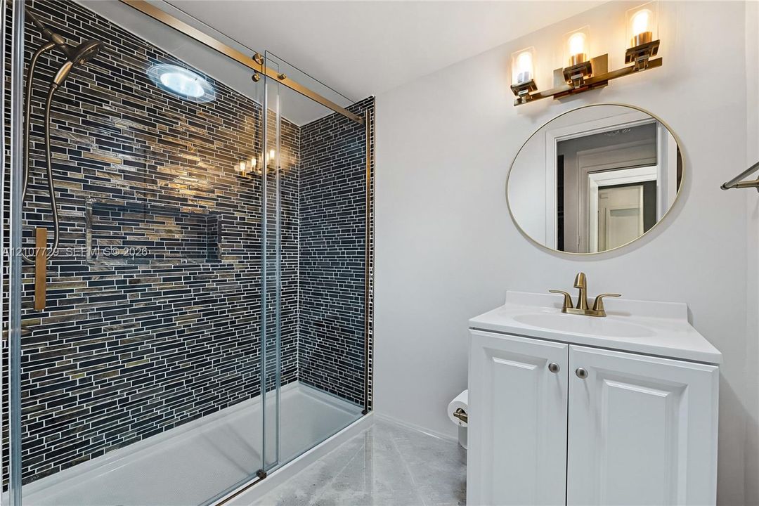 Bathroom featuring a walk-in shower with glass sliding doors and multi-tonal linear tile surround, brass-finish fixtures, white vanity cabinet with an integrated basin, and a circular wall-mounted mirror with a matching brass-finish light fixture
