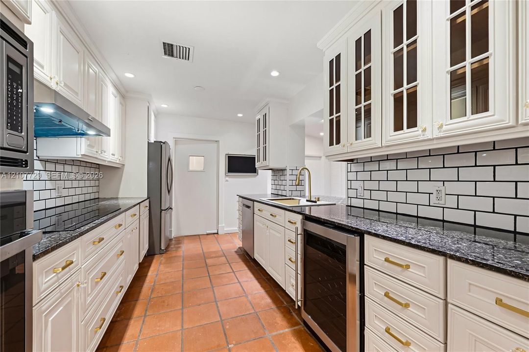 Kitchen featuring white cabinetry with glass-panel upper doors, dark stone countertops, subway tile backsplash, brass-tone hardware, and terra-cotta tile flooring