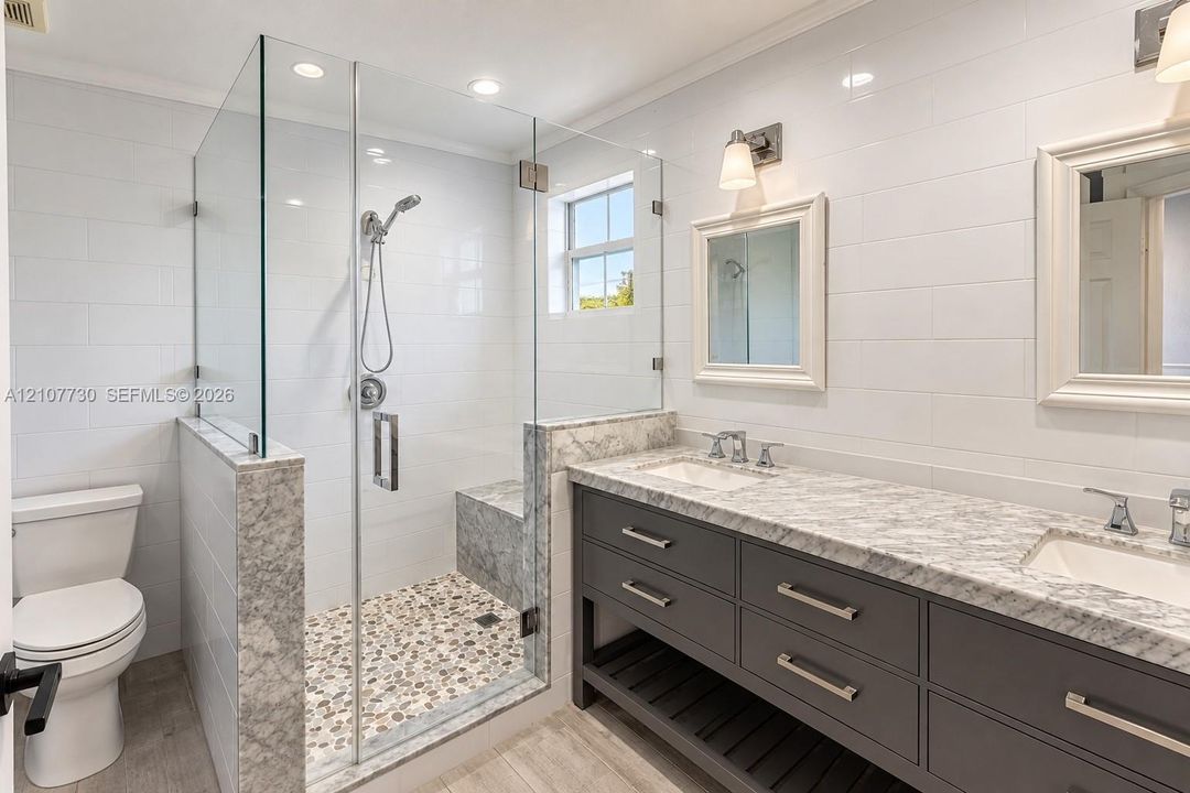 Double vanity with stone countertop, glass-enclosed walk-in shower featuring an integrated bench seat, pebble-style shower flooring, and wall-mounted light fixtures