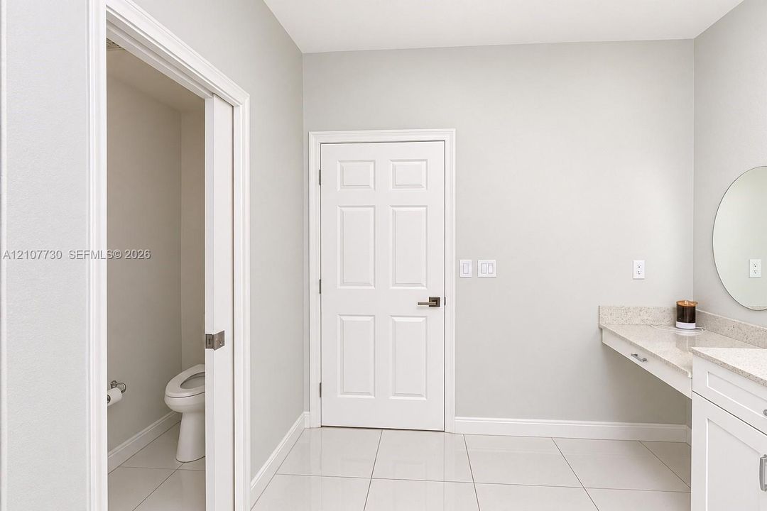 Light-toned tile flooring, white six-panel door, built-in vanity with stone-composite countertop, and wall-mounted circular mirror