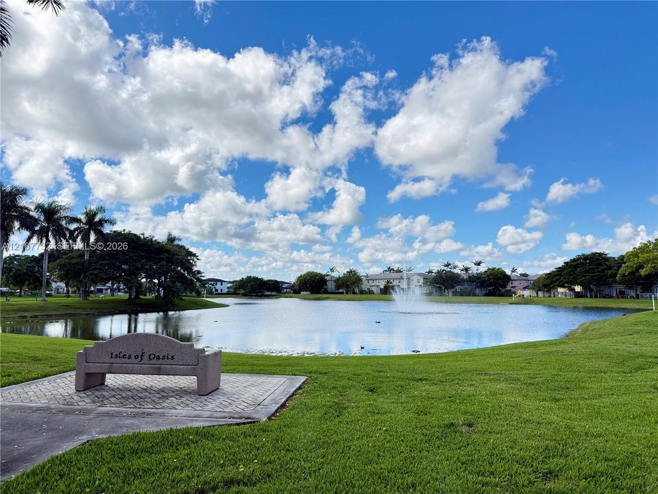 Community pond featuring a central fountain, manicured lawn, stone-finish bench, and patterned paver surround